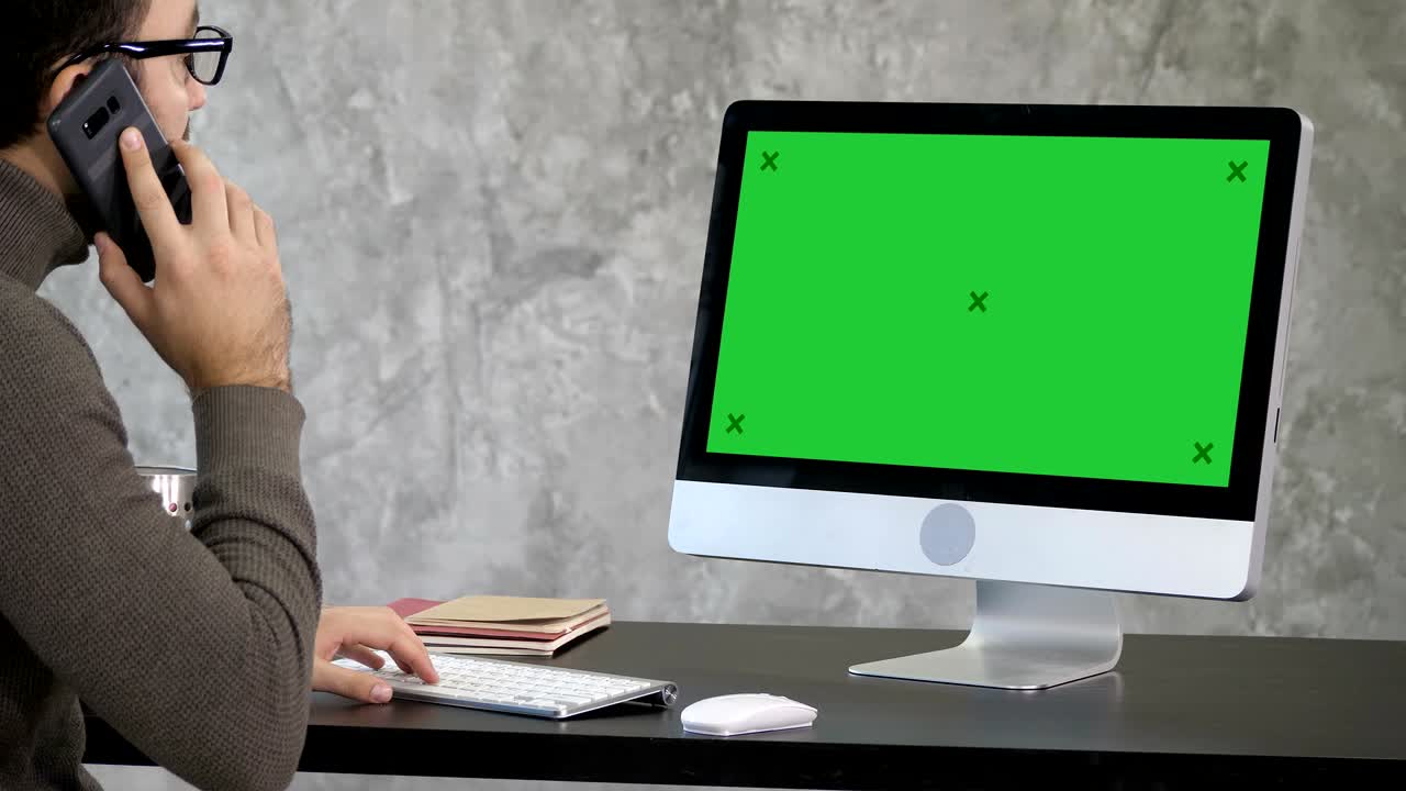 심각한 집중된 남성 사무실 직원은 그의 전화로 전화하고 컴퓨터에서 일합니다. 녹색 화면 모의 디스플레이