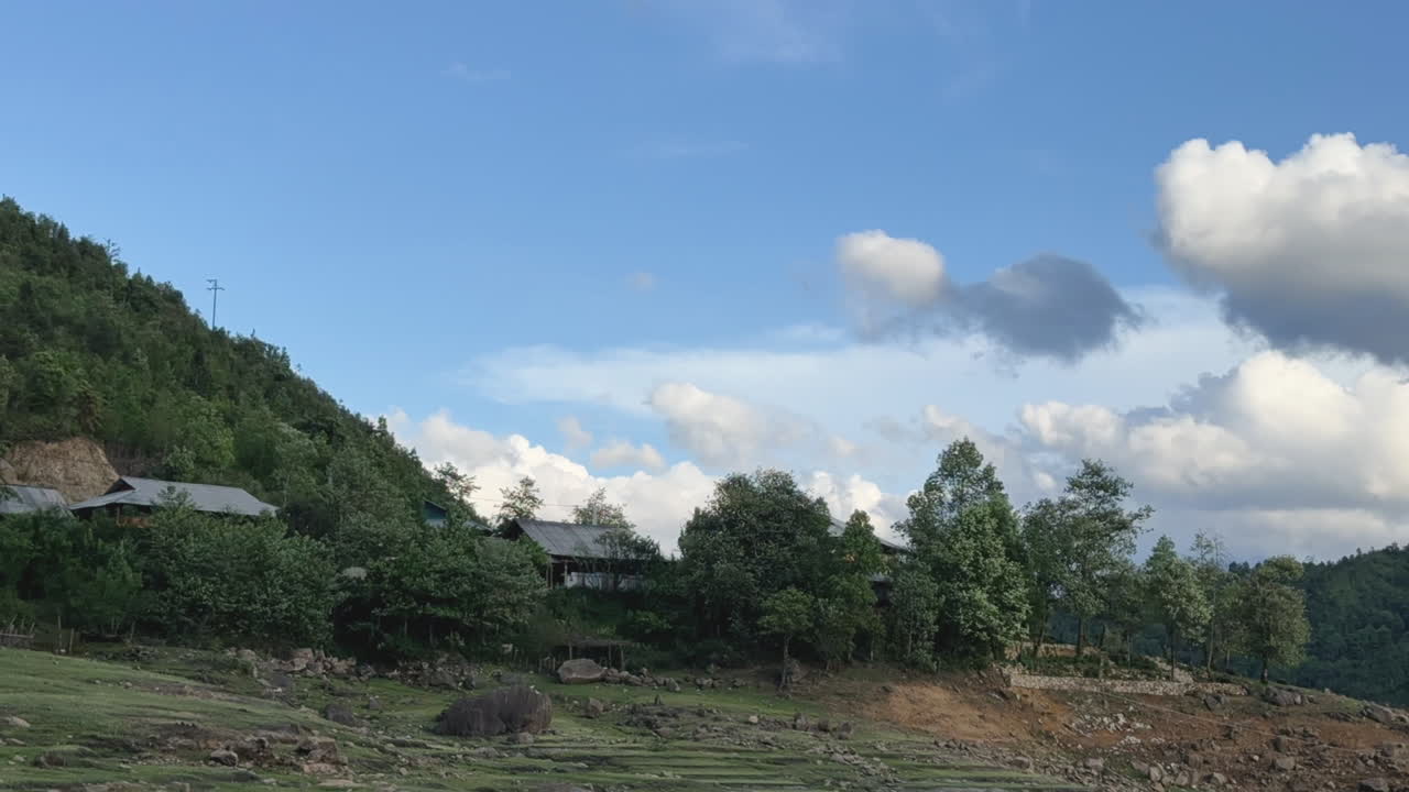 Cloud shadows sweep over a rural landscape with trees and small houses. Fixed shot captures shifting light under a blue sky.