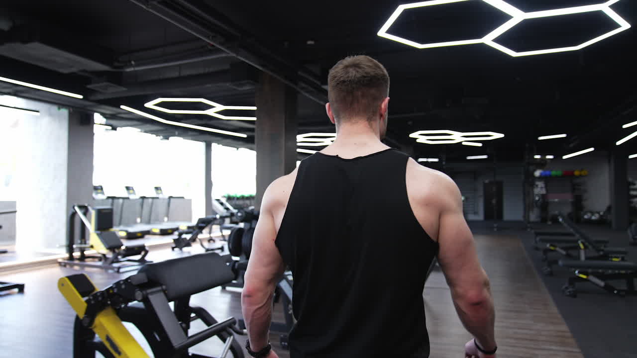 Athletic man walking in a modern fitness center. Strong man coming to a gym to do workout to a simulator. Back view. Slow motion.