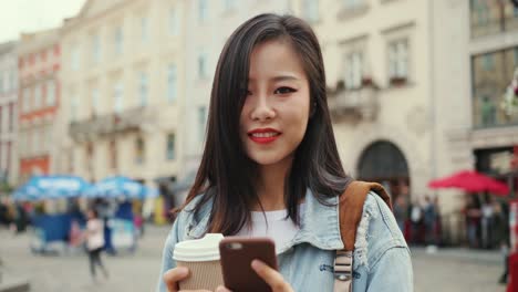 Portrait-Of-The-Beautiful-Young-Woman-Typing-A-Message-And-Chatting-On-The-Smartphone-With-A-Coffee-To-Go-In-Hand-And-Smiling-To-The-Camera-In-The-Town-On-The-Street