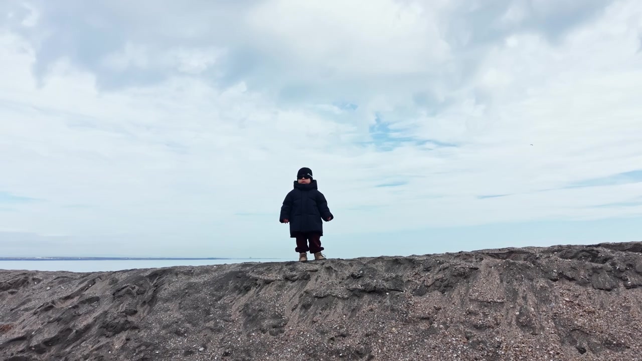 Menino de 2 anos com jaqueta e chapéu de inverno caminha em uma praia de areia. Criança brinca à beira-mar no inverno. Roupas de inverno para uma criança. Passeio de inverno de um menino à beira-mar