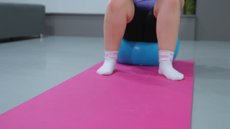 close-up of person balancing on blue exercise ball, sitting and rolling backward on pink mat while holding blue balloon, white socks with pink details visible