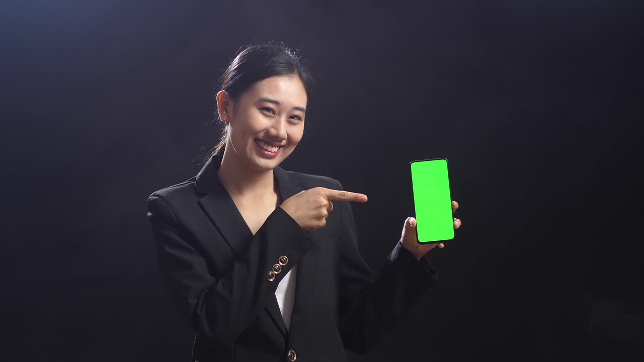 비즈니스 슈트를 입은 아시아 여성 스피커가 블랙 스크린 스튜디오에서 녹색 스크린 스마트폰을 들고 가리키고 있습니다.