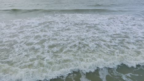 Aerial-birdseye-view-of-big-stormy-waves-breaking-against-the-white-sand-beach,-overcast-day,-seashore-dunes-damaged-by-waves,-coastal-erosion,-climate-changes,-wide-low-drone-shot