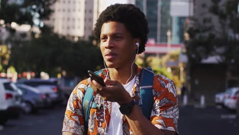 Happy-african-american-man-in-city,-wearing-earphones,-talking-on-smartphone-in-the-street