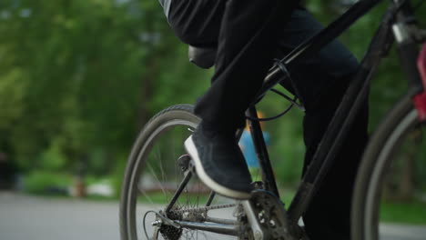 young boy riding a bicycle steadily through a park area, wearing glasses and a grey top, the background includes lush view of trees, an urban building, and blurred people walking in the background