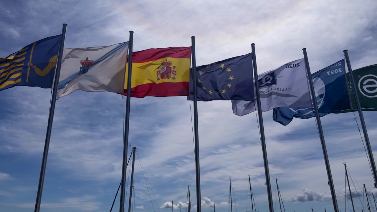 Spanish Galician and European Union flags flying on poles above marina masts under cloudy sky