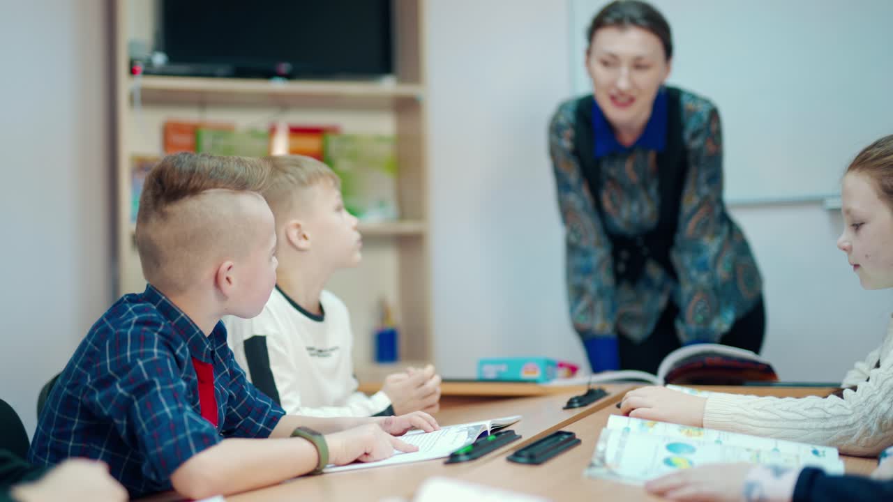 Learning process at primary school. Children sitting at a table in the classroom. Female teacher speaking with young students. Education at school.