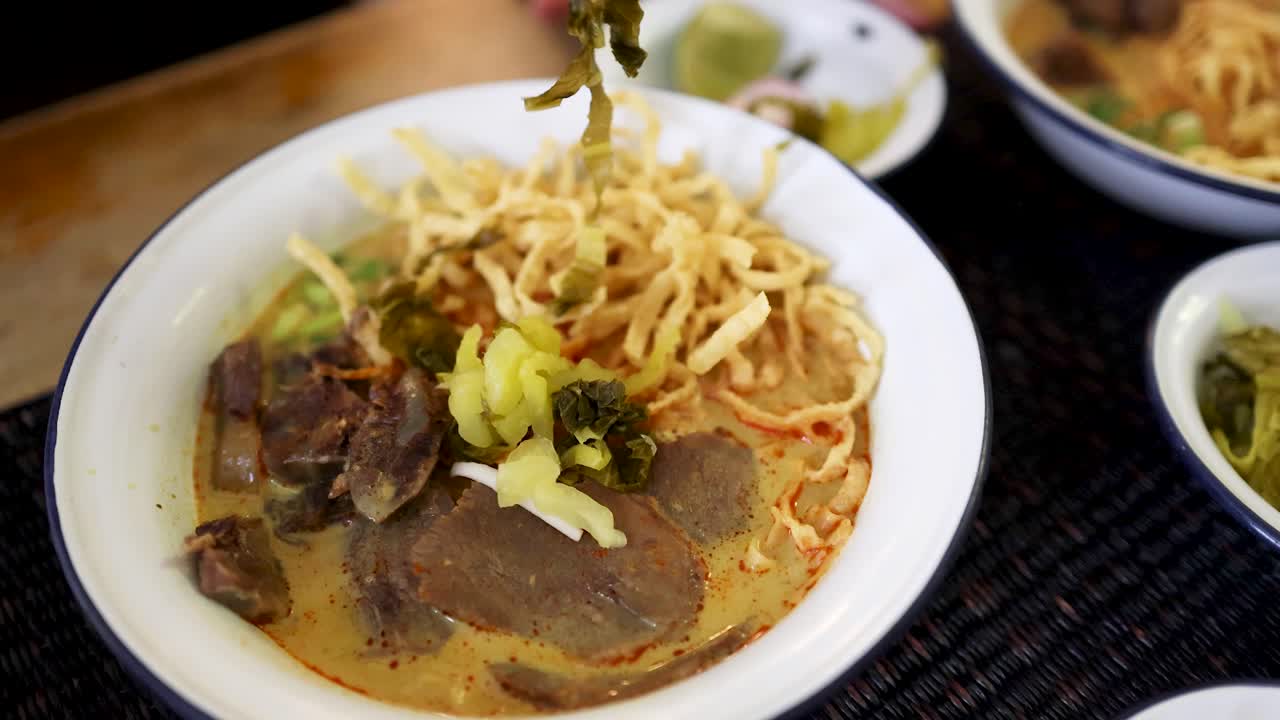 Hand garnishes a bowl of beef curry noodles with pickled mustard greens in a well-lit indoor setting, highlighting traditional Northern Thai cuisine and vibrant textures