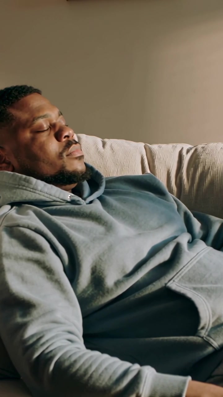 Vertical video: Camera pulling back showing man napping on couch at home with hoodie sweats cushion
