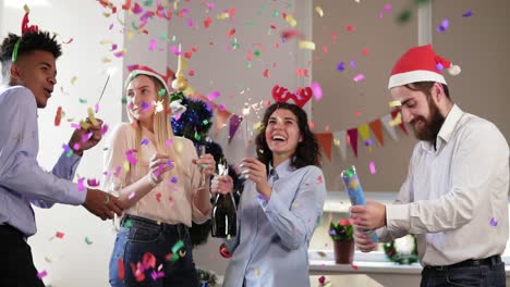 happy multi-ethnic people in funny hats lighting sparklers celebrating christmas and new year in the office, excited diverse