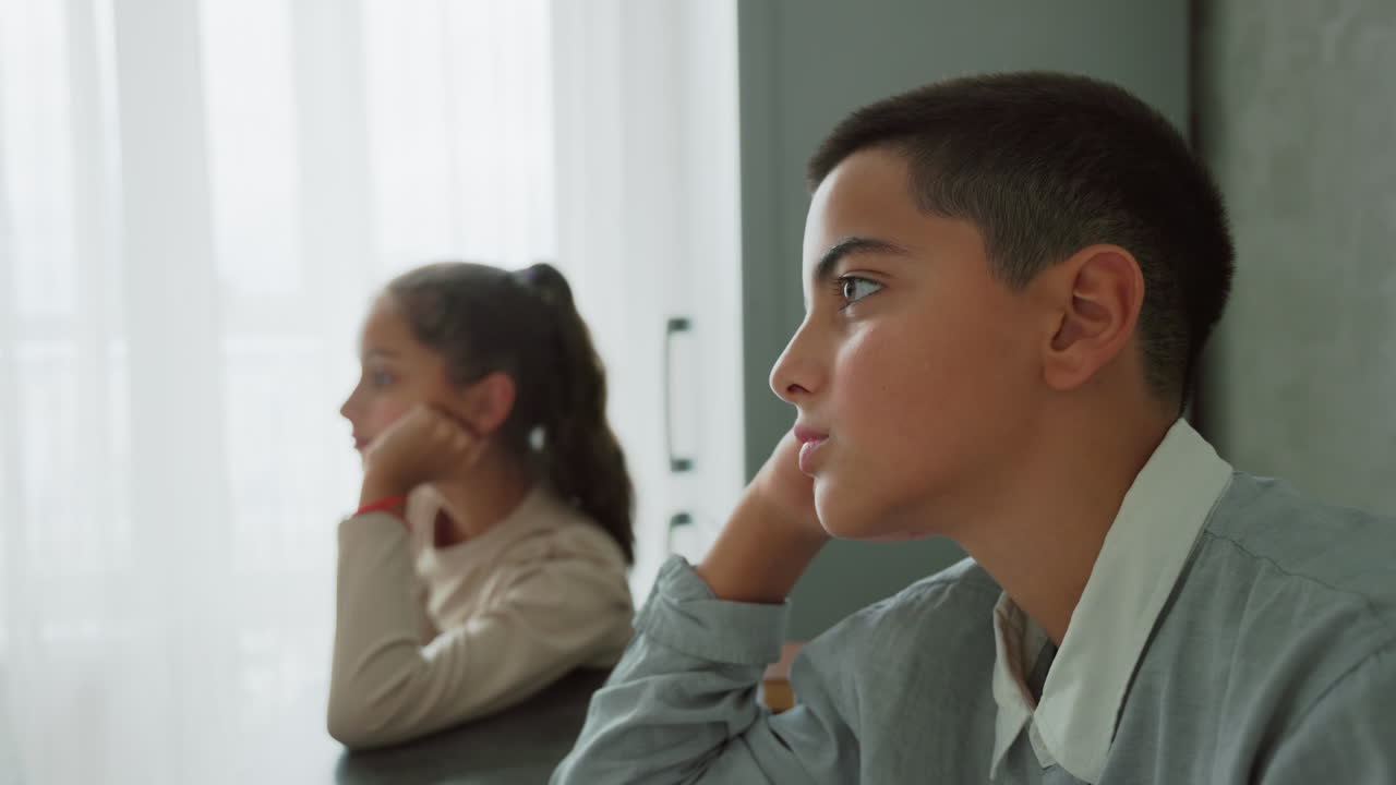boy and girl in school uniforms sitting side by side at table in quiet morning light looking thoughtful and focused while waiting indoors near sheer window curtain in calm domestic atmosphere