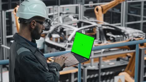 ingeniero de fábrica de automóviles en uniforme de trabajo usando una computadora portátil con pantalla verde. trabajando con software en una instalación de fabricación industrial automotriz dedicada a la producción de vehículos.