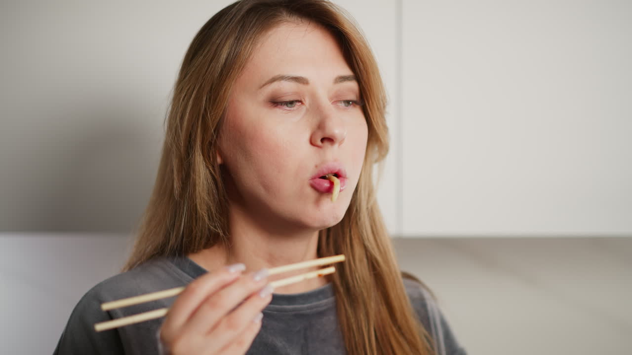 Portrait view of woman with long hair eating noodles with chopsticks while standing in bright kitchen, enjoying light homemade meal in casual indoor setting