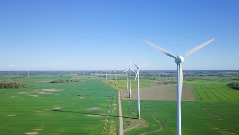 Aerial-view-of-wind-turbines-generating-renewable-energy-in-the-wind-farm,-sunny-spring-day,-low-flyover-over-green-agricultural-cereal-fields,-countryside-roads,-wide-angle-drone-shot-moving-forward