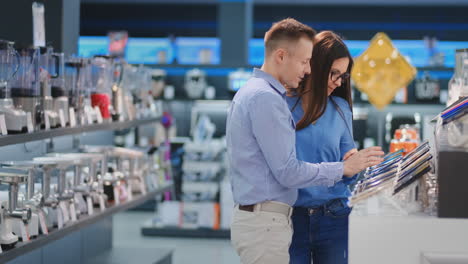 Feliz-Pareja-Familiar,-Hombre-Y-Mujer-Parados-En-El-Mostrador-Con-Teléfonos-Móviles-Vestidos-Con-Ropa-Informal,-Eligiendo-Un-Nuevo-Teléfono-Inteligente-En-Una-Tienda-De-Electrónica-Moderna