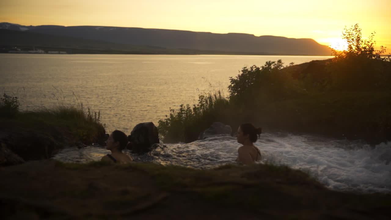 Two women enjoy geothermal hot spring pool at top of waterfall overlooking fjord and ocean at sunset, slow motion