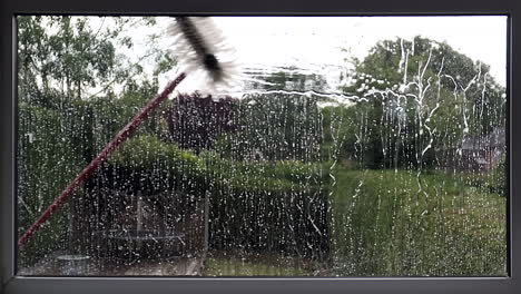 A-window-cleaners-brush-on-a-long-pole-being-use-to-spray-water-to-clean-an-exterior-window-on-the-first-floor-of-a-house