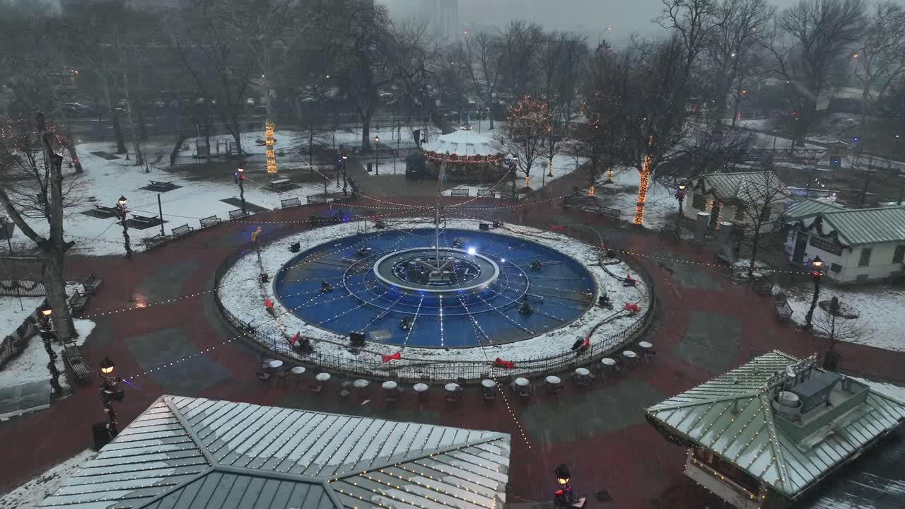Ilmakuva dronella Franklin Squaresta Philadelphiassa talvisen lumisateen aikana. Historiallinen suihkulähde ja puiston polut ovat kevyesti lumen peitossa samalla kun joulunvalot loistavat kaupungin puistossa.