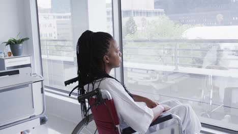 african american female patient in wheelchair looking out window in hospital room, slow motion