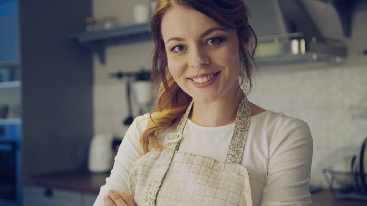 Close up of the beautiful woman in the apron and with a flour on her face posing in the kitchen while cooking and crossing her hands. Portrait. Indoors