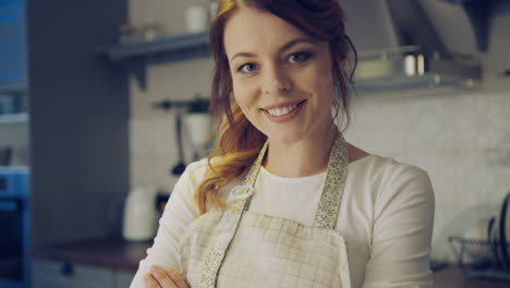 Close-up-of-the-beautiful-woman-in-the-apron-and-with-a-flour-on-her-face-posing-in-the-kitchen-while-cooking-and-crossing-her-hands.-Portrait.-Indoors