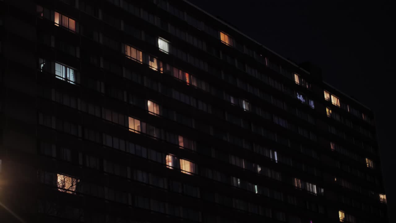 Slow-motion view of an urban residential building at night with illuminated windows seen from a moving car