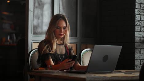 young woman sitting in coffee shop at wooden table, drinking coffee and using smartphone.on table is laptop. girl browsing internet, chatting, blogging. female holding phone and looking on his screen.