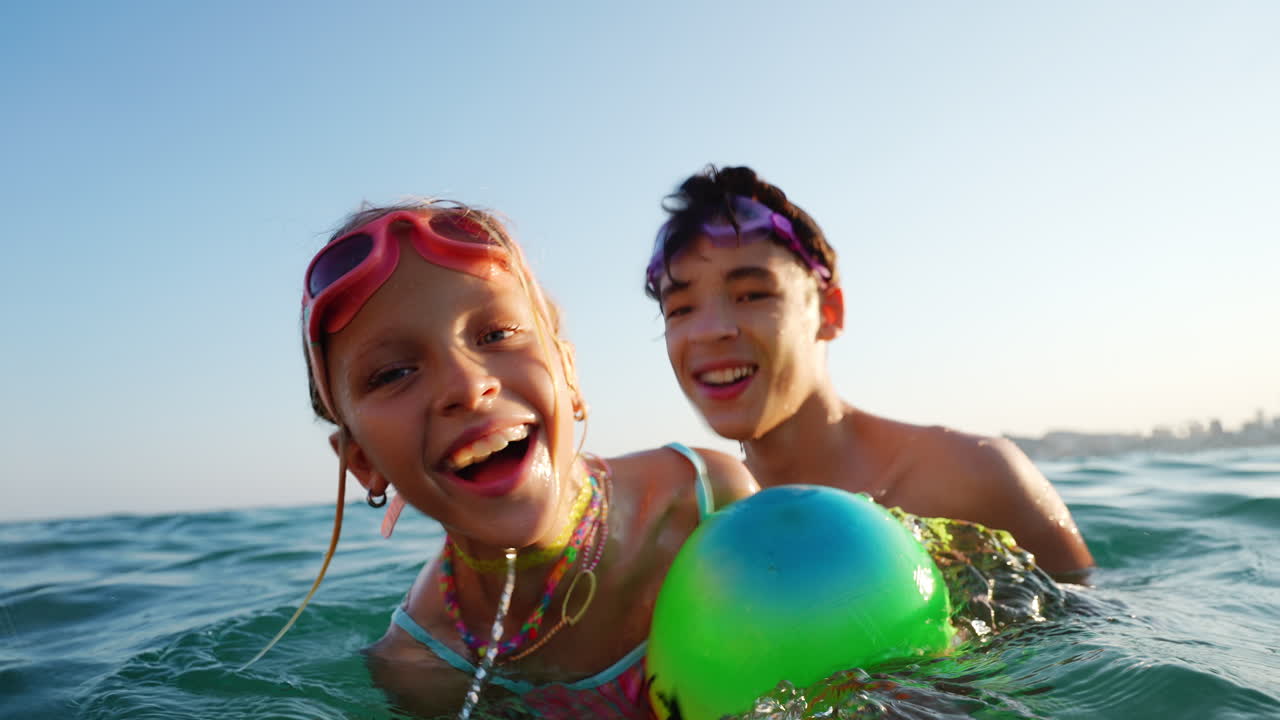 Cheerful siblings wearing swimming goggles enjoying playful moments with colorful inflatable ball, splashing and laughing while experiencing ocean vacation under bright sunlight