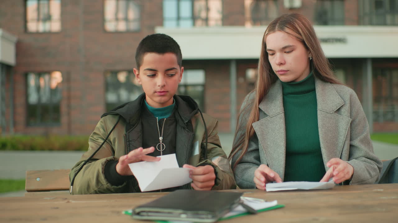 Mother shows young boy how to make paper craft while seated outdoors at wooden table, both focused on folding paper together, highlighting family bonding, learning