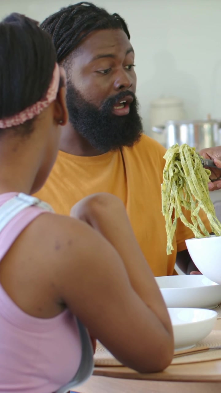 Vertical video: Pointing partner, man in shirt using tongs, serving pasta at kitchen table to eat