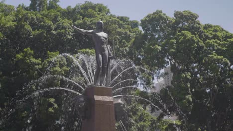 Estatua-De-Un-Hombre-En-Medio-De-Una-Fuente-Con-El-Cielo-Azul-Y-El-Parque-Al-Fondo