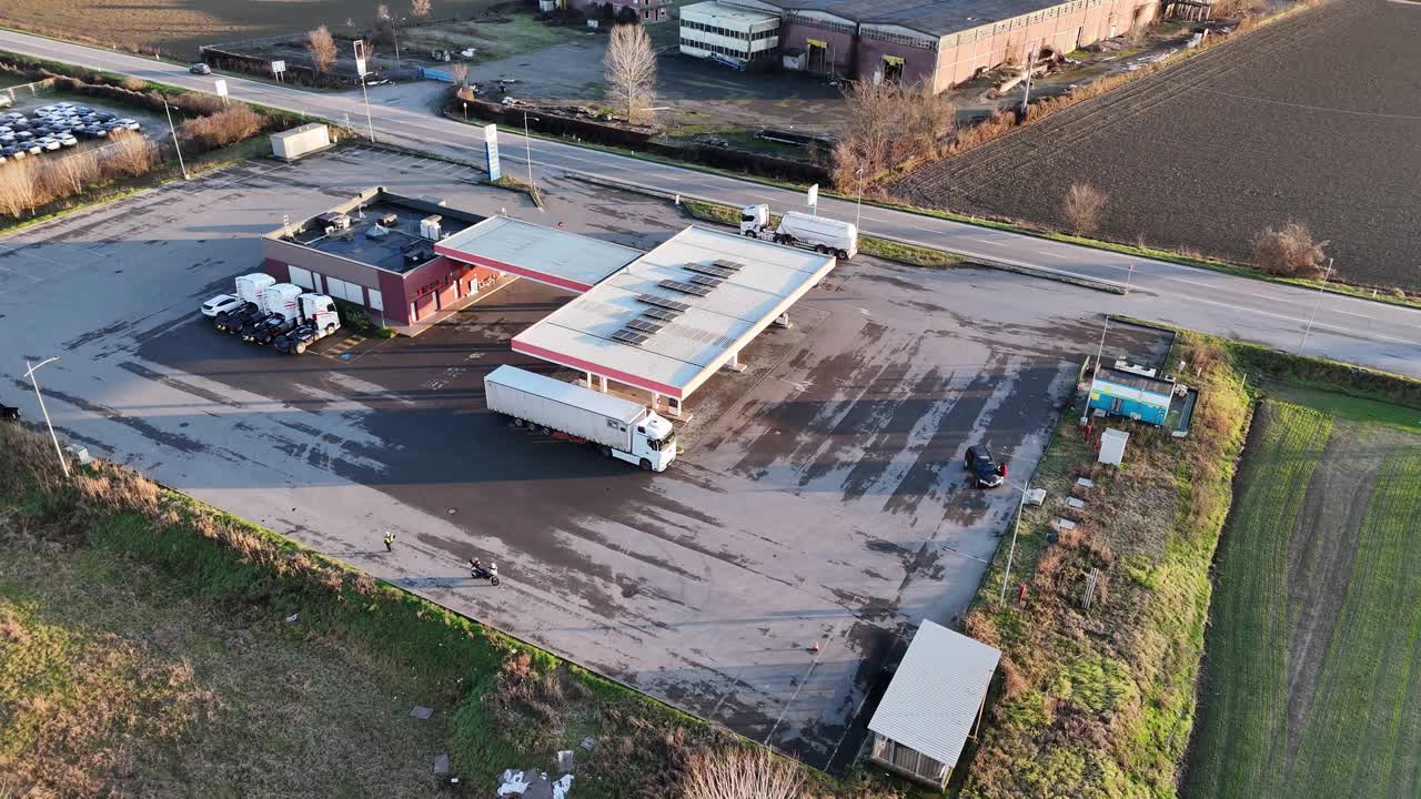 erial drone view shows a gas station along SS10 Strada Padana Inferiore in Piacenza Province, Emilia-Romagna, highlighting fuel pumps, service building, parking area, road access, and flat rural farml