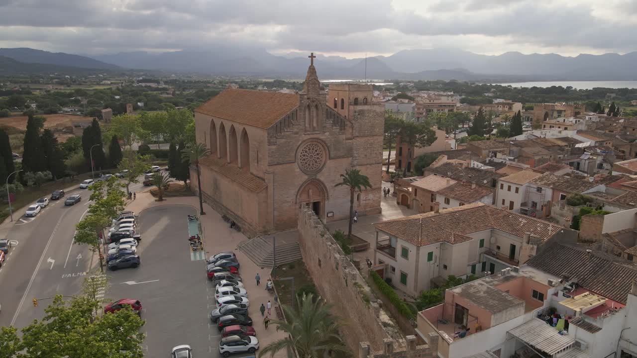 Aerial view of a historic church in Alcúdia, a charming old town on the Spanish island of Mallorca. The church is surrounded by medieval walls, narrow streets, and traditional Mediterranean houses.