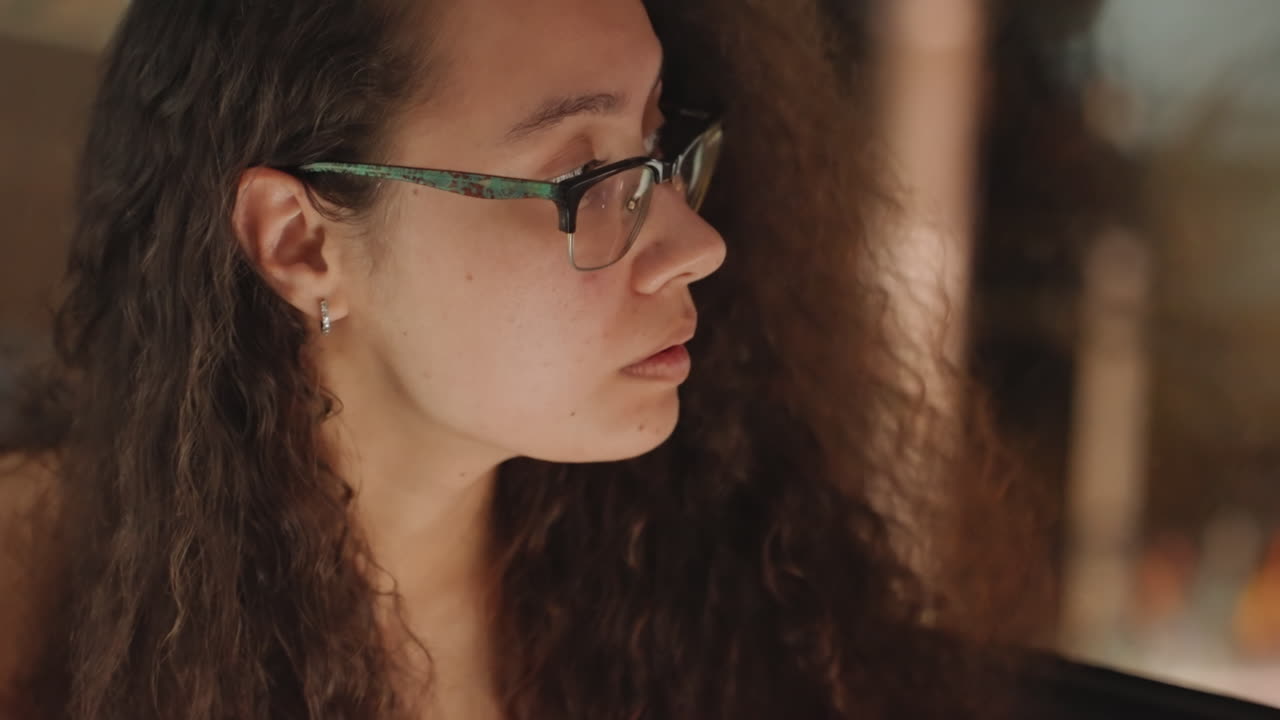 Huésped en primer plano en habitación de hotel estudiando detalles de reserva bajo una lámpara, con un brillo cálido en su cabello rizado y gafas, leyendo documentos y un cuaderno, expresión pensativa, ambiente nocturno tranquilo con una luz tenue.