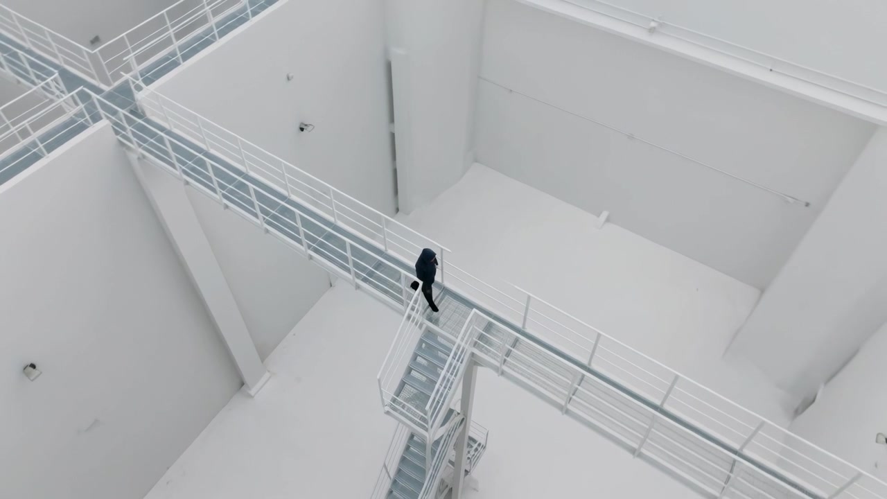 Uomo con cappotto scuro che cammina da un passaggio in una stanza, superando un pianerottolo verso il lato opposto.