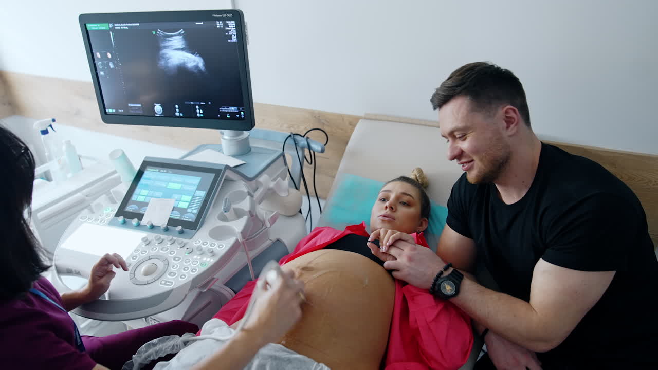 Caucasian expecting parents is in doctor's cabinet. Pregnant woman is checked on ultrasound machine. Smiling man holds his wife's hand.