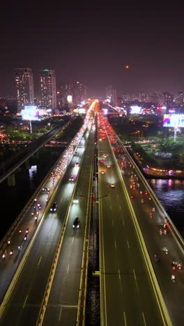 vertical aerial night view of heavy traffic on a major bridge over the saigon river in ho chi minh city, vietnam, illuminated by city lights and billboard signs.