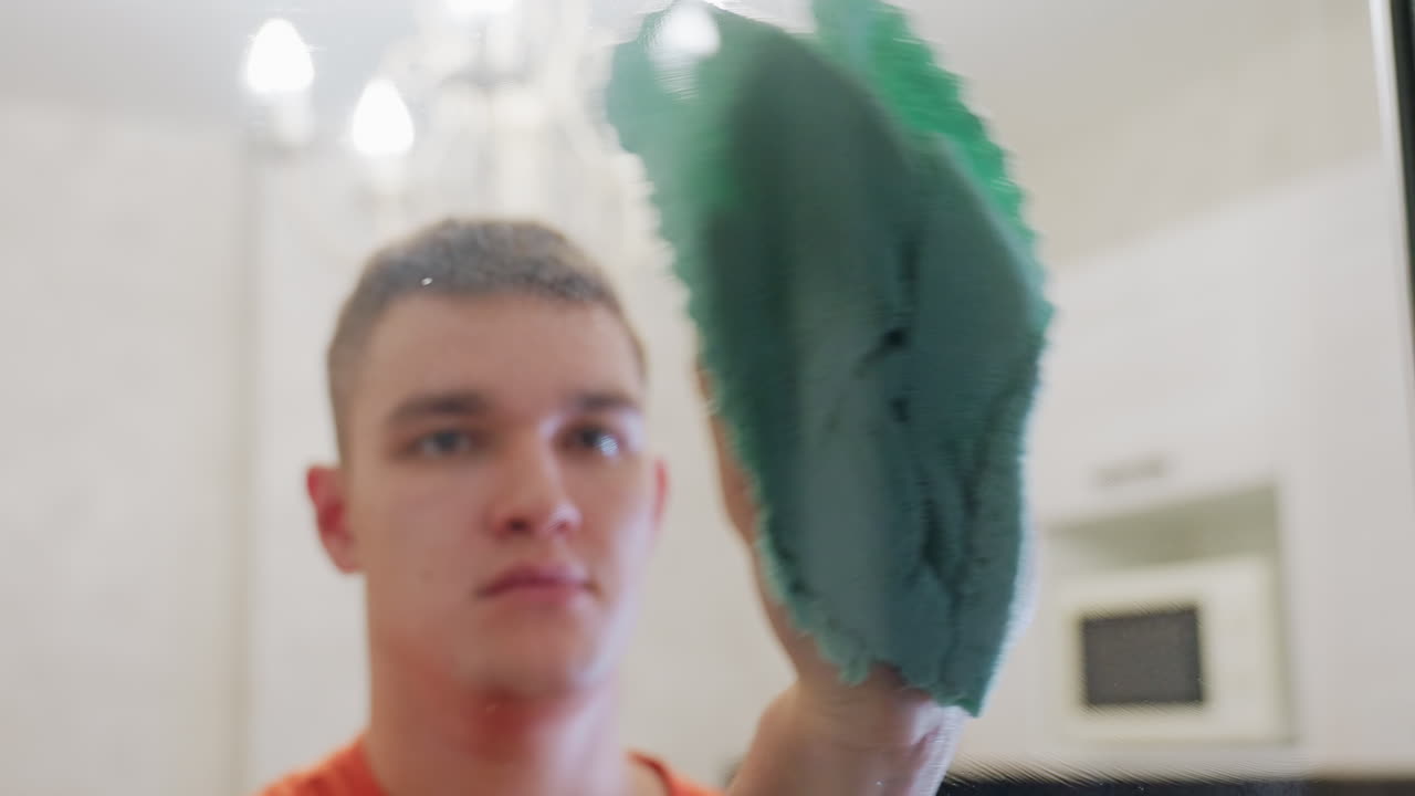 Man in orange shirt cleaning mirror with green towel, carefully wiping away water streaks and spray marks, focusing on neatness, cleanliness, household hygiene and daily maintenance routine indoors