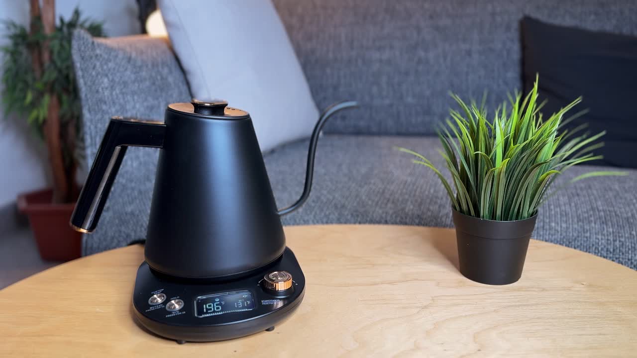 Primo piano di una mano che solleva un bollitore elettrico a collo d'oca nero opaco dalla sua base su un tavolo di legno all'interno, con divano e pianta in vaso in una luce soffusa diurna, mentre si preparano bevande calde.