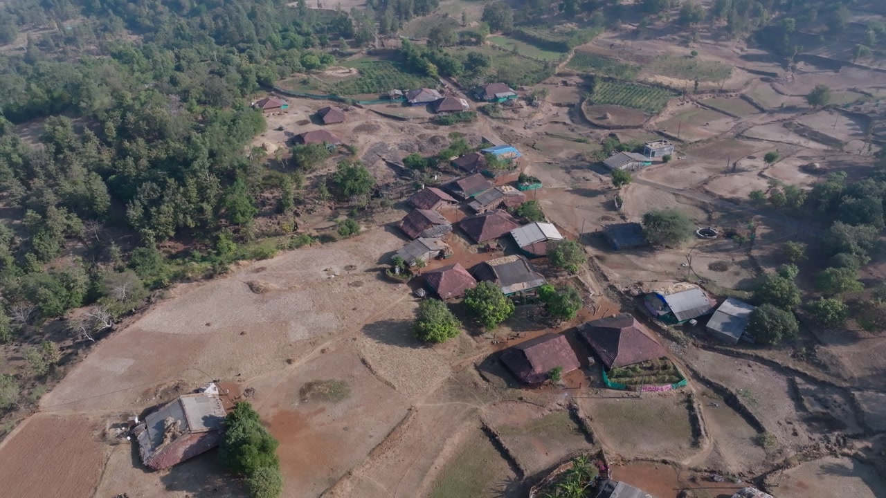 Aerial drone footage pulling back over a rural Indian village, revealing traditional clay-roofed homes, dirt pathways, surrounding crop fields, and forested hillsides under diffused daylight.