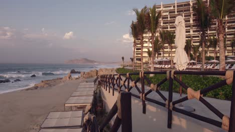 wide shot of a beachfront resort with palm trees and cabanas, focused on the sandy shore and waves crashing on rocks near a modern building.