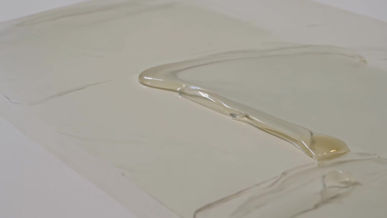 Una goccia si forma sul bordo destro di un liquido viscoso pallido che scorre verso sinistra su un pannello da banco, con un raschietto nelle vicinanze.
