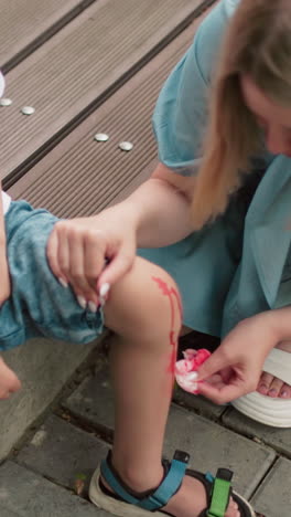 una madre se agacha suavemente y limpia la pierna lesionada de su hijo con un pañuelo mientras otra persona es parcialmente visible en el fondo