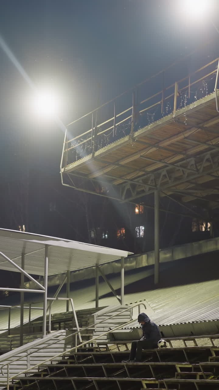 A man dressed in black and wearing a jacket sits alone in an empty stadium at night, with a soccer ball beside him with background illuminated by bright stadium light