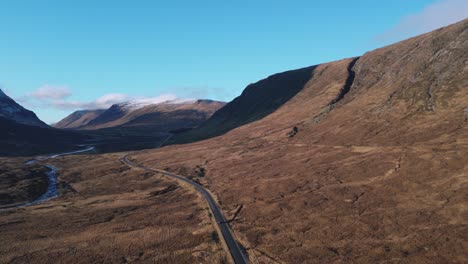 Carretera-Serpenteante-En-El-Valle-De-Glencoe-Con-Montañas-Escarpadas-Y-Cielos-Despejados,-Vista-Aérea