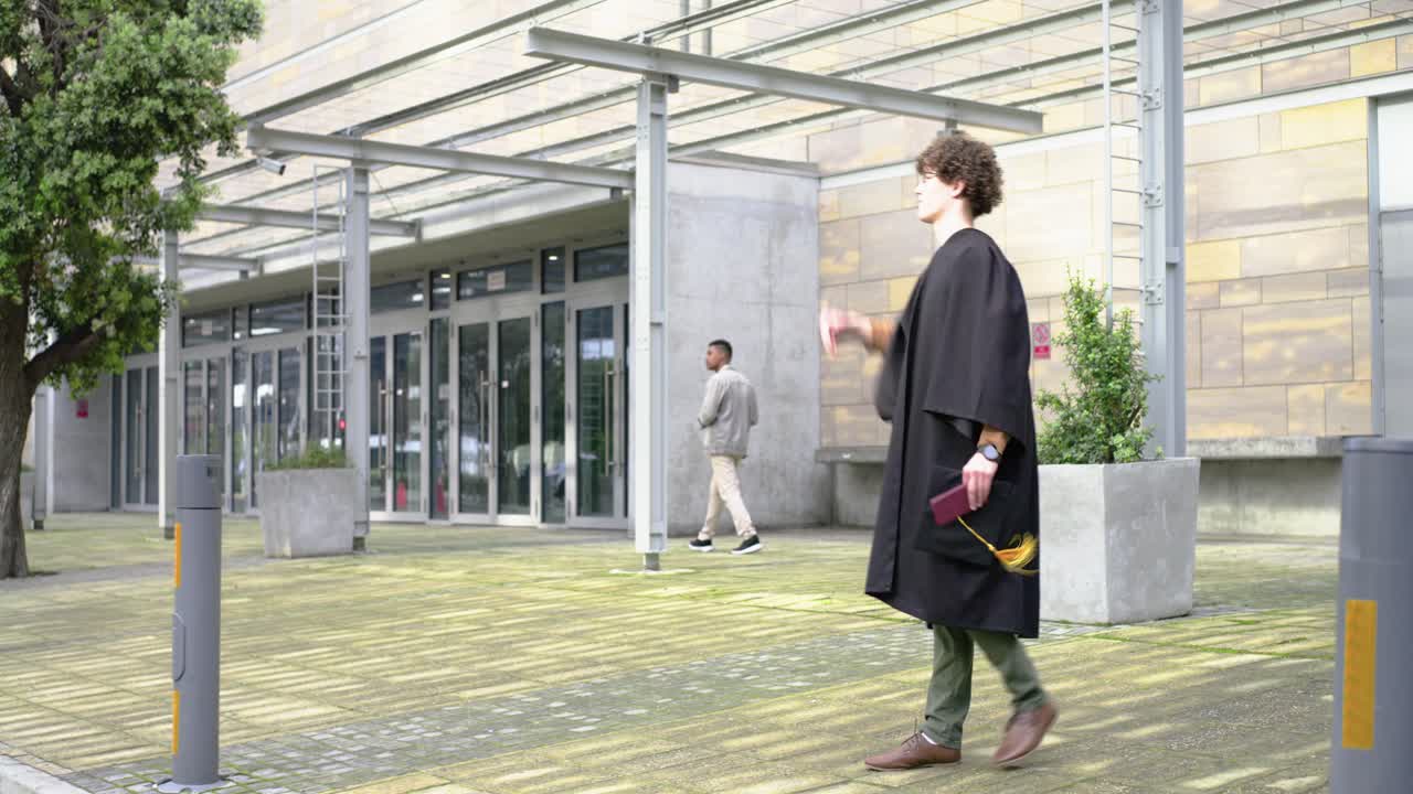Laureato adulto in toga cammina nel campus, con tocco e cartella in mano, saluta un passante ed entra da una porta.