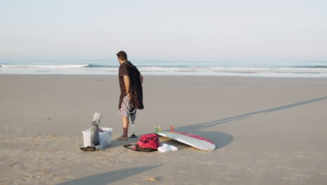 Lange-Aufnahme-Eines-Männlichen-Surfers-Mit-Künstlichem-Bein,-Der-An-Der-Küste-Steht-Und-Sich-Auf-Das-Training-Auf-Dem-Surfbrett-Vorbereitet