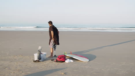 long shot of male surfer with artificial leg standing on coastline, preparing for training on surfboard