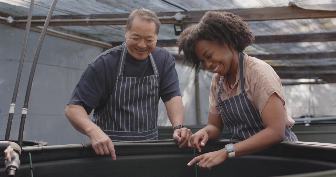 Senior Chinese man aproned pointing at black tank guiding African woman testing water in greenhouse